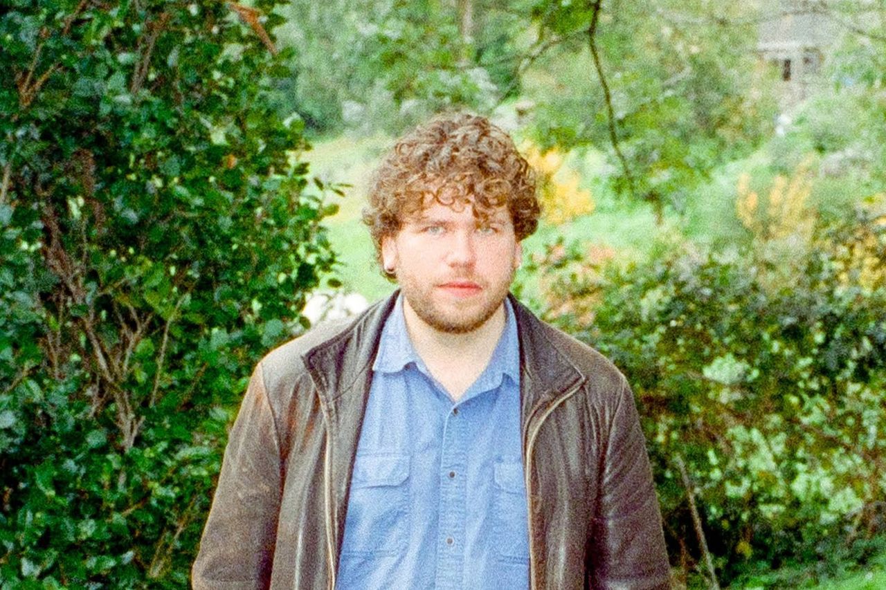 Portrait of musician Ricky Albeck with curly brown hair wearing a brown jacket and blue button-up shirt outdoors in front of green foliage.